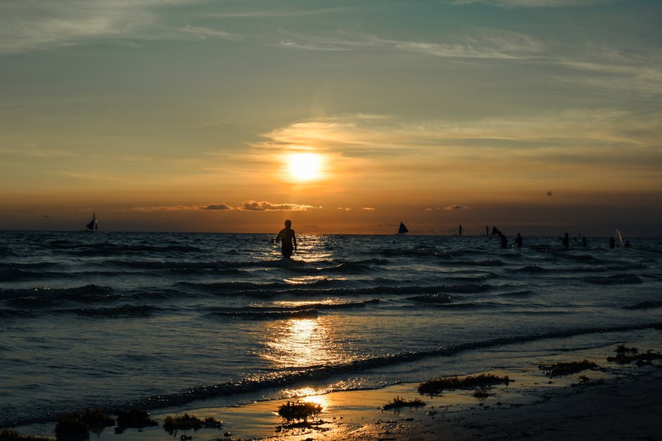 Silhouetted figure at sunset with sailboats and gentle waves in Kalibo, Philippines.