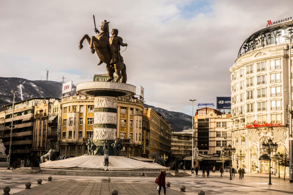 Aged stone sculpture of warrior on horse near house facades and snowy mountain under cloudy sky in town