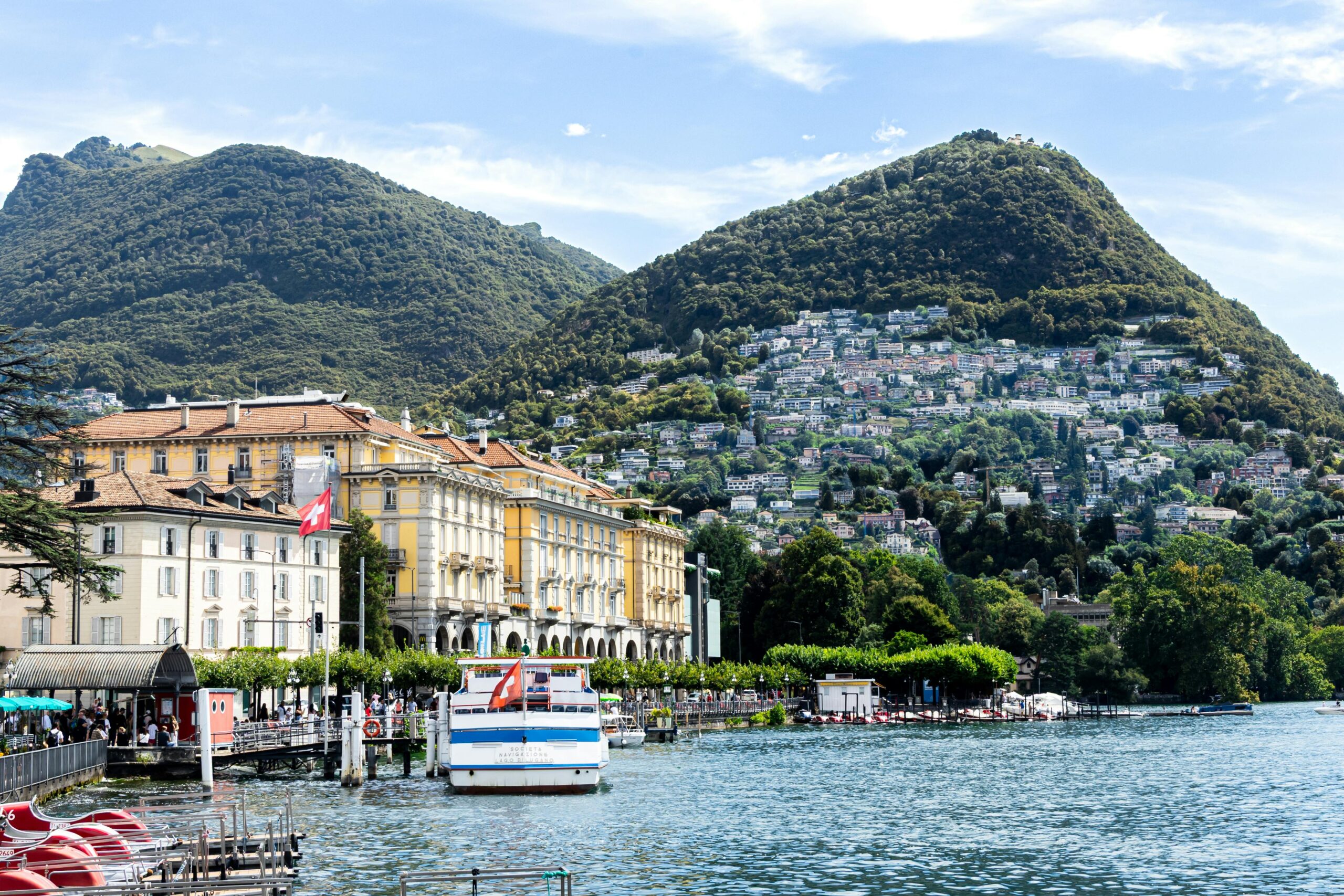 Scenic view of Lake Lugano with mountains and elegant buildings against a bright sky.